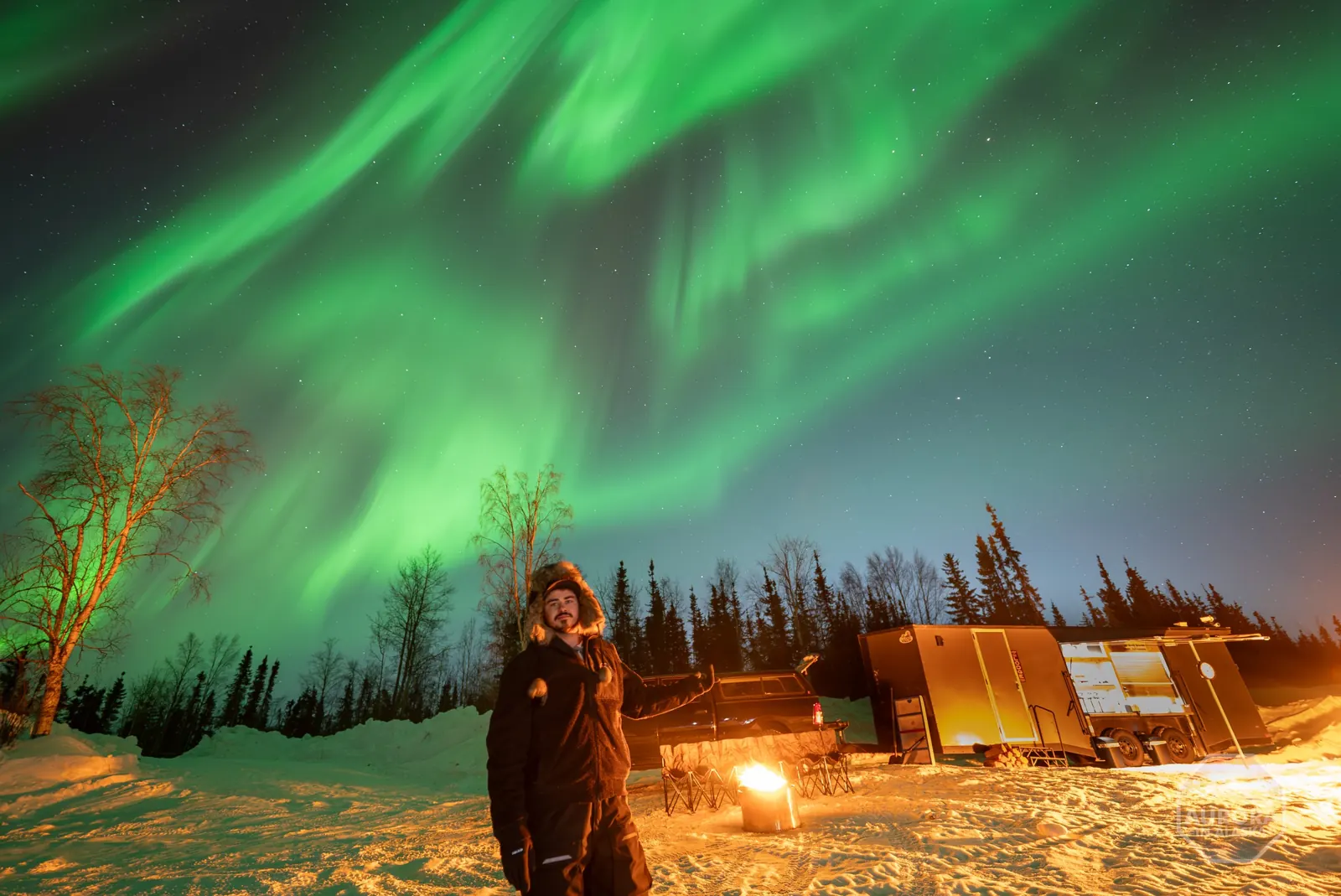 A SkyChase guide by the fire, with the glowing Mobile Lodge behind and aurora overhead, February 25, 2025.
